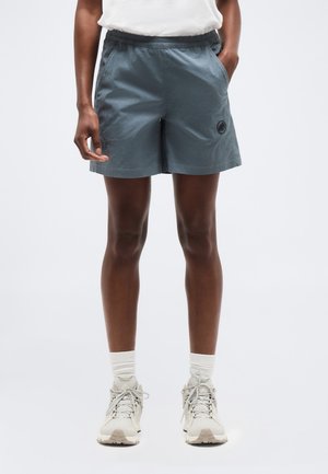 Personne portant un short gris à taille élastique avec poches latérales et des baskets beige clair avec des chaussettes blanches sur un fond uni.