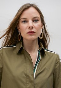 Olive green shirt with a buttoned collar, featuring a striped pattern in dark green and white, and an embroidered heart detail on the chest.