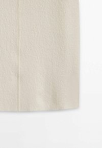 Gros plan sur un tissu en laine beige avec une couture visible et une surface texturée, montrant la partie inférieure d'un vêtement sur fond blanc.