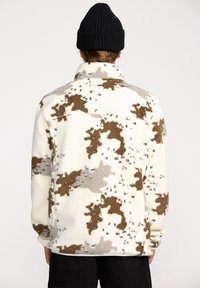 Veste en polaire blanche avec des motifs abstraits marron et gris, dotée d'un col montant et d'une coupe décontractée. Texture douce et manches longues.