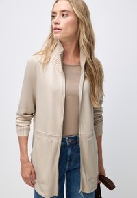 Cardigan zippé beige avec col à côtes, poches latérales et manches longues, porté par-dessus un haut taupe clair et associé à un jean bleu.