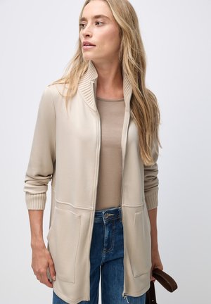 Cardigan zippé beige avec col à côtes, poches latérales et manches longues, porté par-dessus un haut taupe clair et associé à un jean bleu.