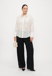 White sequined button-up shirt with long sleeves, paired with black wide-leg trousers and a beige handbag. The shirt has a classic collar.