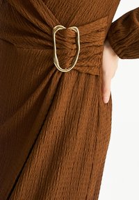 Brown textured dress featuring a twisted design with a large metallic oval buckle. Fabric appears crinkled with a soft finish.