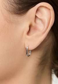 Srebrna kolczyk w kształcie obręczy na przekłutym uchu. Projekt ma gładką, błyszczącą powierzchnię z subtelną fakturą i małym błyszczącym akcentem.