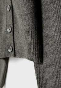 Tmavomodrý kardigán vyrobený z rebrového úpletu, s okrúhlymi čiernymi gombíkmi a voľne strihaným spodným lemom. Textúra vyzerá mäkko a útulne.