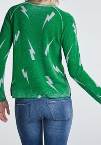 Pull vert avec des motifs d'éclairs blancs, à manches longues et à coupe décontractée, assorti avec un jean en denim bleu.