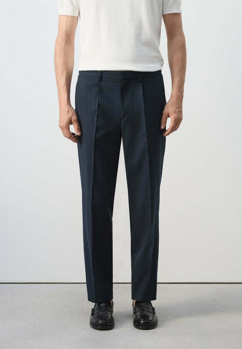 Homme portant un pantalon de costume noir, des chaussures en cuir noires et une chemise blanche à manches courtes, debout devant un fond clair uni.