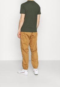 Olivově zelené tričko, krátké rukávy, kulatý výstřih; spárováno s krémovými tapered kalhotami, elastické manžety, a bílými teniskami s červenými akcenty.