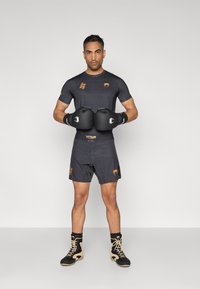 Μαύρο αθλητικό κοντομάνικο μπλουζάκι με διακοσμητικό σχέδιο και πορτοκαλί λεπτομέρειες, μαύρα γάντια πυγμαχίας, μαύρο σορτς και παπούτσια ψηλού τύπου.