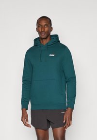 Felpa con cappuccio di un color teal scuro con tasca frontale, caratterizzata da un piccolo logo Puma bianco e arancione sul petto. Tessuto in cotone misto strutturato.