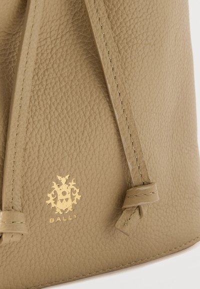 Κοντινή λήψη από μπεζ υφή δέρματος με κόμπο κορδονιού και μικρό χρυσό έμβλημα λογότυπου ανάγλυφο πάνω από τη ραμμένη άκρη.