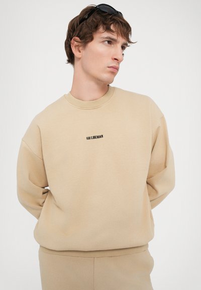 Beige sweatshirt med ribbet halsudskæring og manchetter, prydet med et lille sort logo på brystet. Glat stof og afslappet pasform.