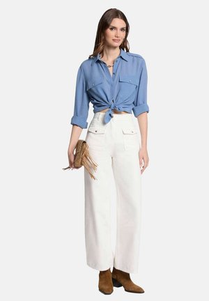 Chemise cropped bleu clair avec un devant noué, associée à un pantalon large blanc. Des bottines brunes et un sac à franges complètent la tenue.