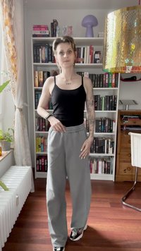 Rövid hajú személy, fekete toppban, szürke nadrágban és tetoválásokkal áll egy könyvespolc előtt egy fa padlós szobában.