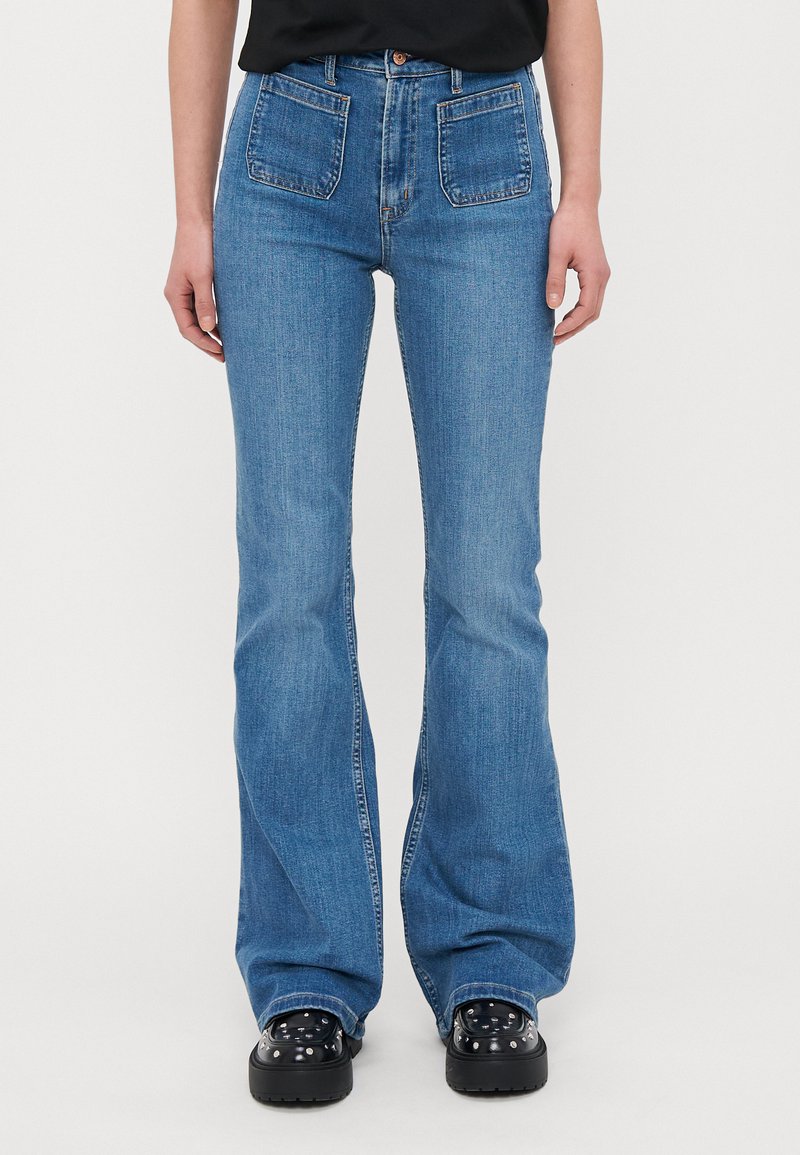 Bredde blå denimjeans med høy midje, fem lommer og små broderidetaljer. Brukt med svarte platåsko.
