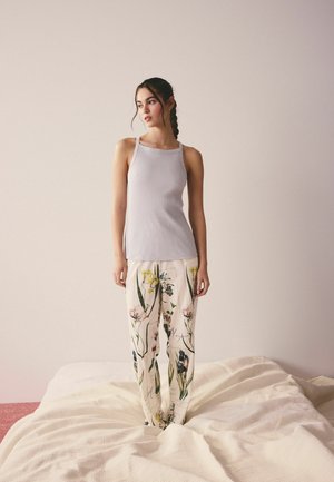 Vrouw met gevlochten haar staat op een bed en draagt een mouwloos lavendelkleurig geribbeld topje en een witte broek met kleurrijke bloemenprints.