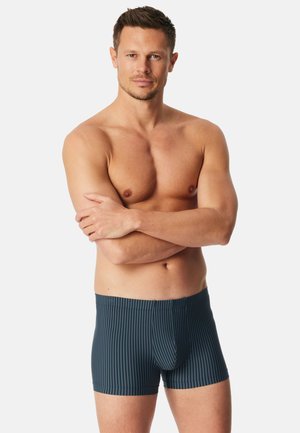 Mann mit kurzen braunen Haaren, verschränkten Armen, trägt blaue gestreifte Boxershorts vor schlichtem weißem Hintergrund.