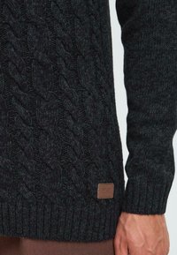 Maglione lavorato a maglia scura caratterizzato da un motivo a trecce, polsini a coste testurizzati e un piccolo etichetta in pelle marrone con la scritta "ISOLID" sul lato.