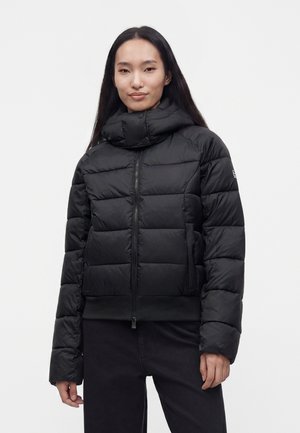 Vrouw met lang donker haar, gekleed in een zwarte doorgestikte donzen jas met een hoge kraag en een ritssluiting aan de voorkant tegen een effen lichte achtergrond.