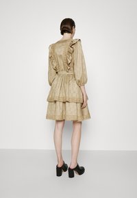 Robe beige à volants, avec un motif hexagonal texturé, épaules froncées, détails en dentelle et taille ceinturée. Portée avec des chaussures à talons carrés noires.