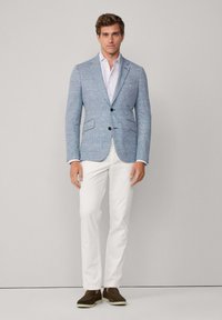 Helles blaues strukturiertes Blazer mit zwei Knöpfen, kombiniert mit einem weißen Hemd und schmalen weißen Hosen. Braune Slipper runden das Outfit ab.