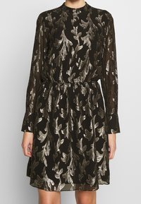 Robe noire à manches longues, ornée de motifs de feuilles métalliques dorées, avec une taille cintrée et une texture de tissu douce et transparente.