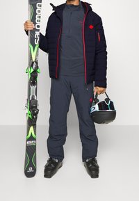 Skiutrustning som består av en marinblå puffjacka med röda detaljer, grå baslager, marinblå byxor, svarta kängor, skidor med gröna detaljer och en hjälm.