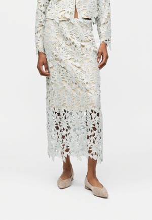 Vrouw draagt een beige onderjurk met een lichtblauwe bloemenkant midi-rok en een bijpassend kanten jasje met lange mouwen, gecombineerd met beige platte schoenen.