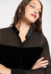 Czarna bluzka zapinana na guziki, wykonana z przezroczystej tkaniny z aksamitną wstawką na piersi, z kołnierzykiem typu mandarynka i gładką fakturą.