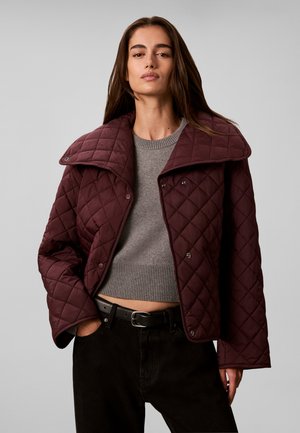 Femme portant une veste matelassée bordeaux par-dessus un pull court gris et un pantalon noir avec une ceinture, debout avec une main dans la poche.