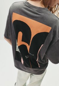 Grå oversized t-shirt med et stort grafisk motiv i orange og sort, med tekstelementer, korte ærmer og rund halsudskæring.
