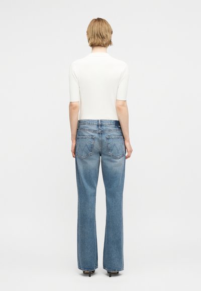 Άτομο που στέκεται με την πλάτη γυρισμένη, φοράει λευκό μπλουζάκι με κοντά μανίκια και ανοιχτό μπλε τζιν παντελόνι κατευθείαν μπατζάκια με ραφές τσέπης και μαύρες γόβες.