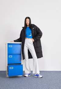 Melna pūka jakas, zils teksturēts īsais tops, svītroti balti plaša kāju bikses un zili un balti sporta apavi, stāvot blakus zilai dokumentu skapim.