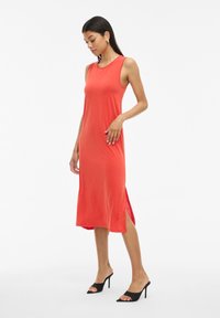 Vestido midi rojo sin mangas en tejido suave con aberturas laterales, acompañado de tacones negros destalonados, que muestra una silueta elegante.