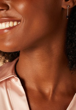 Zwarte kraal hangoorbel met een zilveren ketting die de nek accentueert. Een gladde, glanzende afwerking op de huid en een lichtroze satijnen blouse.