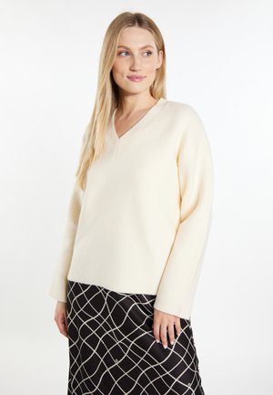 Blonde Frau trägt einen cremefarbenen V-Ausschnitt-Pullover und einen schwarzen Rock mit weißem Kettenglied-Muster, sieht zur Seite mit einem leichten Lächeln.