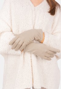 Gants beiges avec une texture lisse et un design à cinq doigts, portés par des bras croisés, sur un pull en tricot blanc doux.