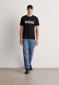 Mann, der einen schwarzen Hut, ein schwarzes T-Shirt mit dem Schriftzug "BOSS", hellblaue Jeans und schwarze Sneakers trägt, vor einem schlichten weißen Hintergrund.