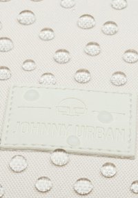 Közelkép egy világos anyagról vízcseppekkel, valamint egy öltött címkéről, amelyen szemüveg ikon és a "Johnny Urban" felirat található.