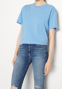 Camiseta de algodón azul claro con cuello redondo y mangas cortas, que presenta un pequeño logo bordado en el pecho, combinada con jeans de mezclilla desgastados.