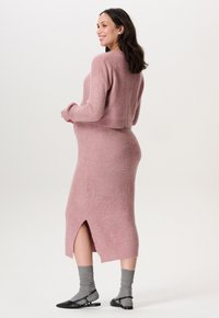 Ensemble en tricot rose comprenant un pull et une jupe à silhouette ajustée, avec des fentes latérales. Complété par des chaussettes grises et des ballerines noires.
