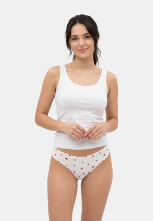 Kobieta ubrana w biały bezrękawnik i białą bieliznę z czerwonymi wzorami serc, stojąca na jednolitym jasnym tle.