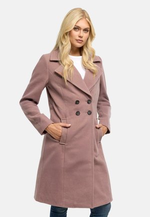 Kvinde med blondt hår iført en mauve dobbeltradet frakke med hænderne i lommerne mod en ensfarvet lys baggrund.