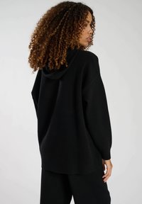 Svart hoodie med avslappnad passform, med en stor huva, ribbade muddar och en mjuk textur, visad från baksidan.