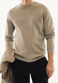 Beige svetr z bavlněné směsi s dlouhými rukávy, kulatým výstřihem, vroubkovanou texturou a mírně zakřiveným spodním lemem, doplněný béžovou bundou.