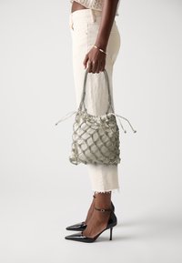 Sac en tissu vert et doux avec un motif en losanges texturé et des accents argentés, doté d'une fermeture à cordon et de poignées tressées, tenu à bout de bras.