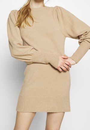 Woman wearing a beige long-sleeve knit sweater dress with ribbed cuffs and hem, standing with arms crossed at the waist.