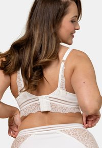 Soutien-gorge en dentelle blanche avec des bretelles réglables, présentant une bande en dentelle texturée, une fermeture à crochets et une finition en voile contrastante.
