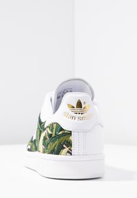 Valkoinen tennari, jossa vihreä ja vaaleanpunainen kirjailtu lehvistökuvio sivulla, kultainen "stan smith"-brändäys ja harmaa sisävuori.